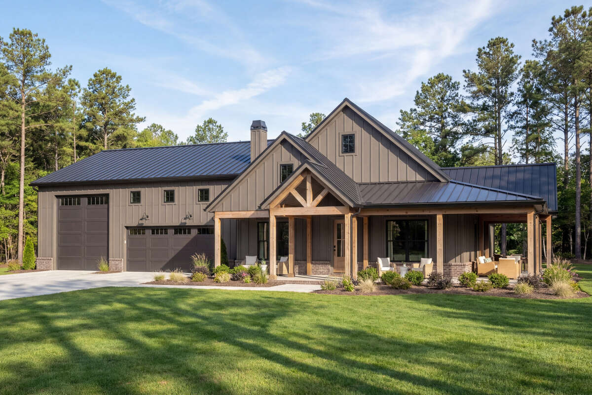 Front exterior rendering of Barndominium Plan 009-00317 showing a single-story Shouse with a wrap-around covered porch, board and batten siding, rustic wood garage doors on a 3-car RV-bay garage, and a brick perimeter skirt