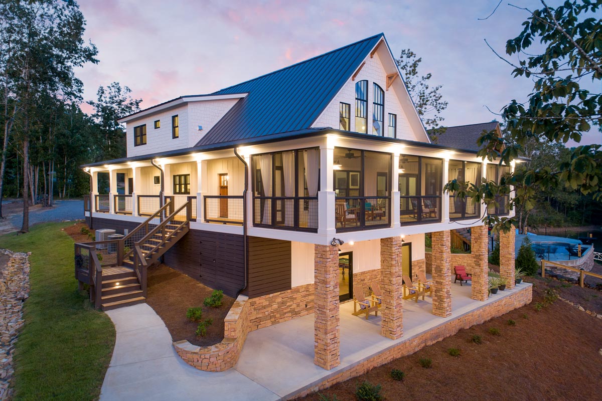 Exterior rendering of Architectural Designs Plan 18302BE, a two-story Craftsman lake house with a three-sided wraparound porch, screened view-side section, metal roof, stacked stone pillars, and gabled dormers set on a sloping lot